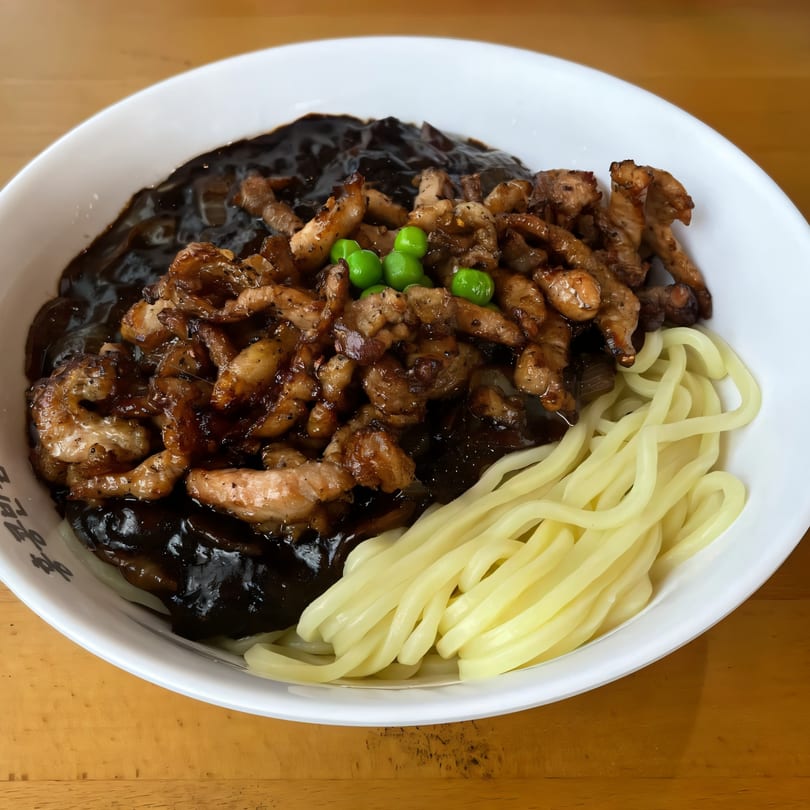 A white bowl of Paik's Noodle Cypress jajangmyeon with thick noodles, black bean sauce, pork, and peas.