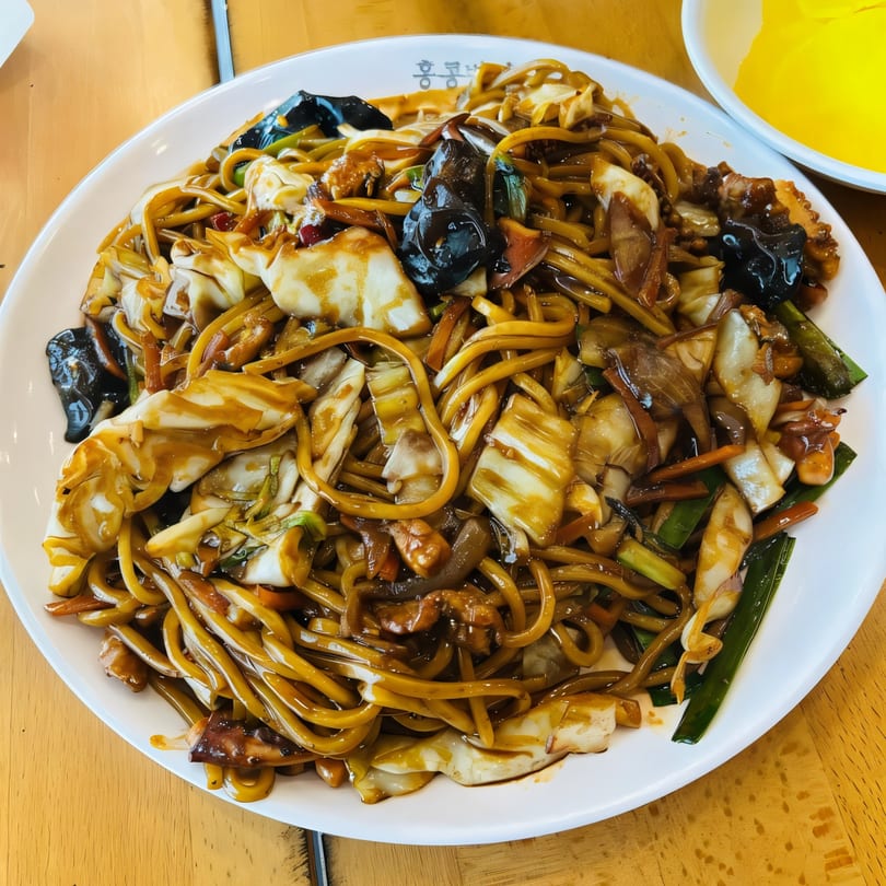 A large plate of Korean Jajangmyeon noodles with dark sauce, vegetables, and seafood.